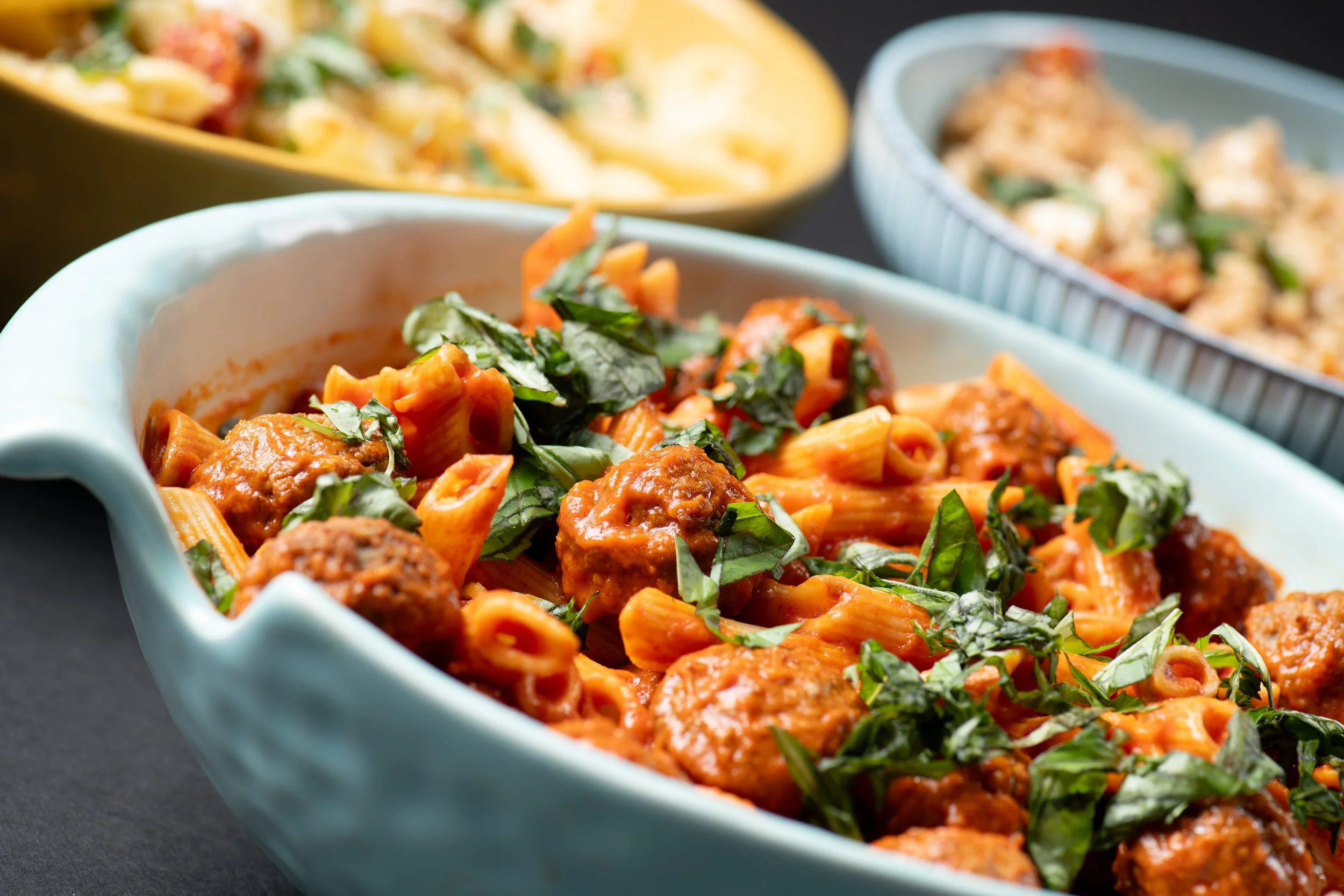 Close-up of a dish with penne pasta, meatballs, and tomato sauce topped with fresh basil, with other pasta dishes in the background.