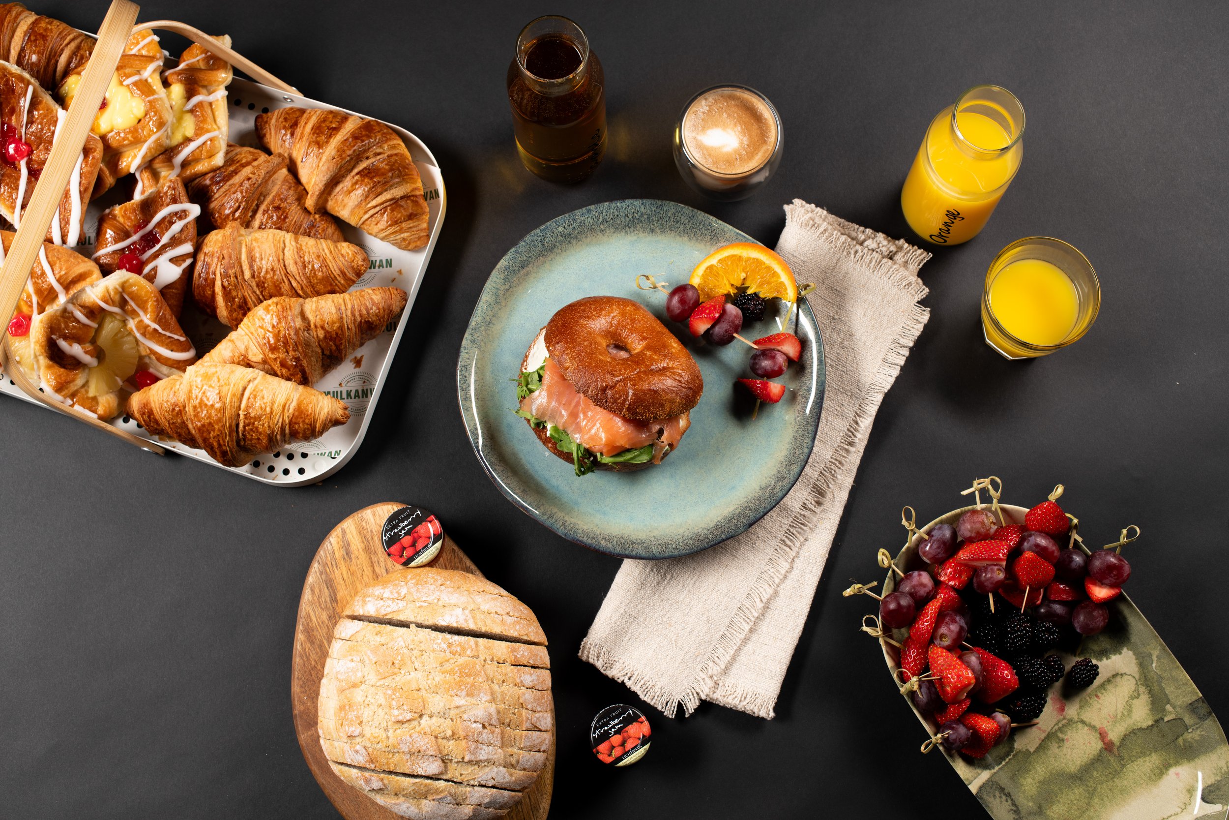 Assorted breakfast spread with croissants, bagel, fruits, and juice on a dark surface