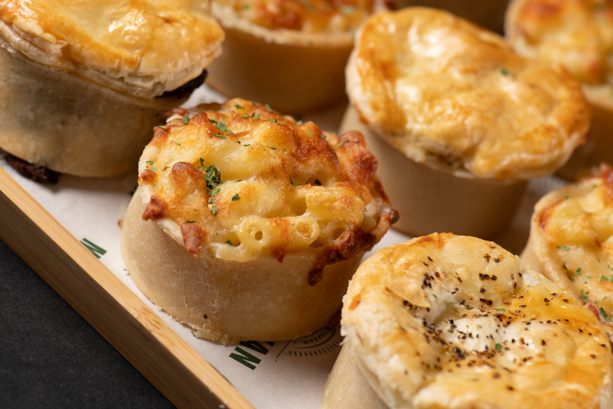 A close-up of assorted savory pies, including a mac and cheese pie topped with herbs, displayed on a wooden tray.
