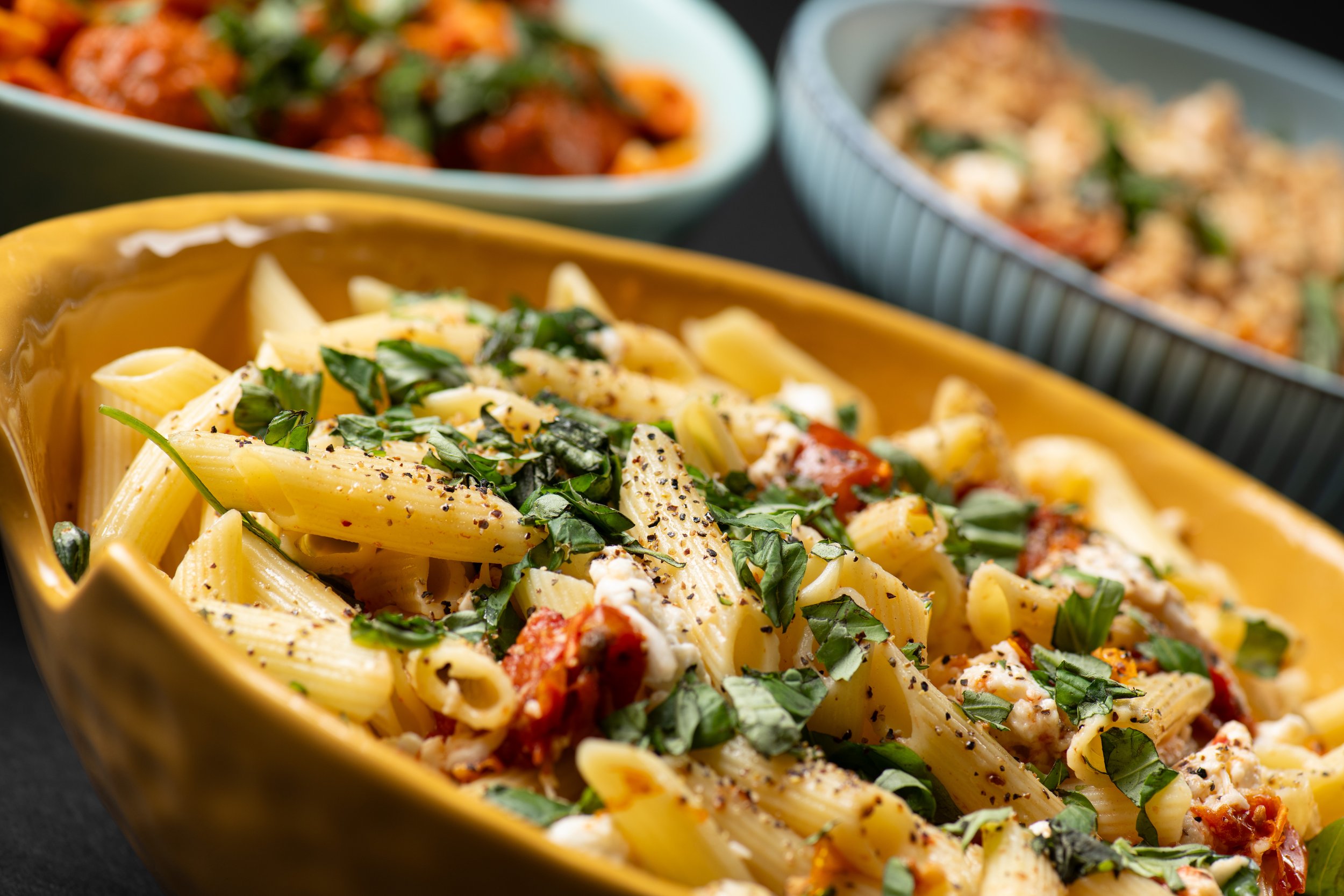 Close-up of a yellow bowl filled with penne pasta topped with fresh basil, cracked pepper, tomatoes, and cheese. Two other bowls with various dishes in the background.