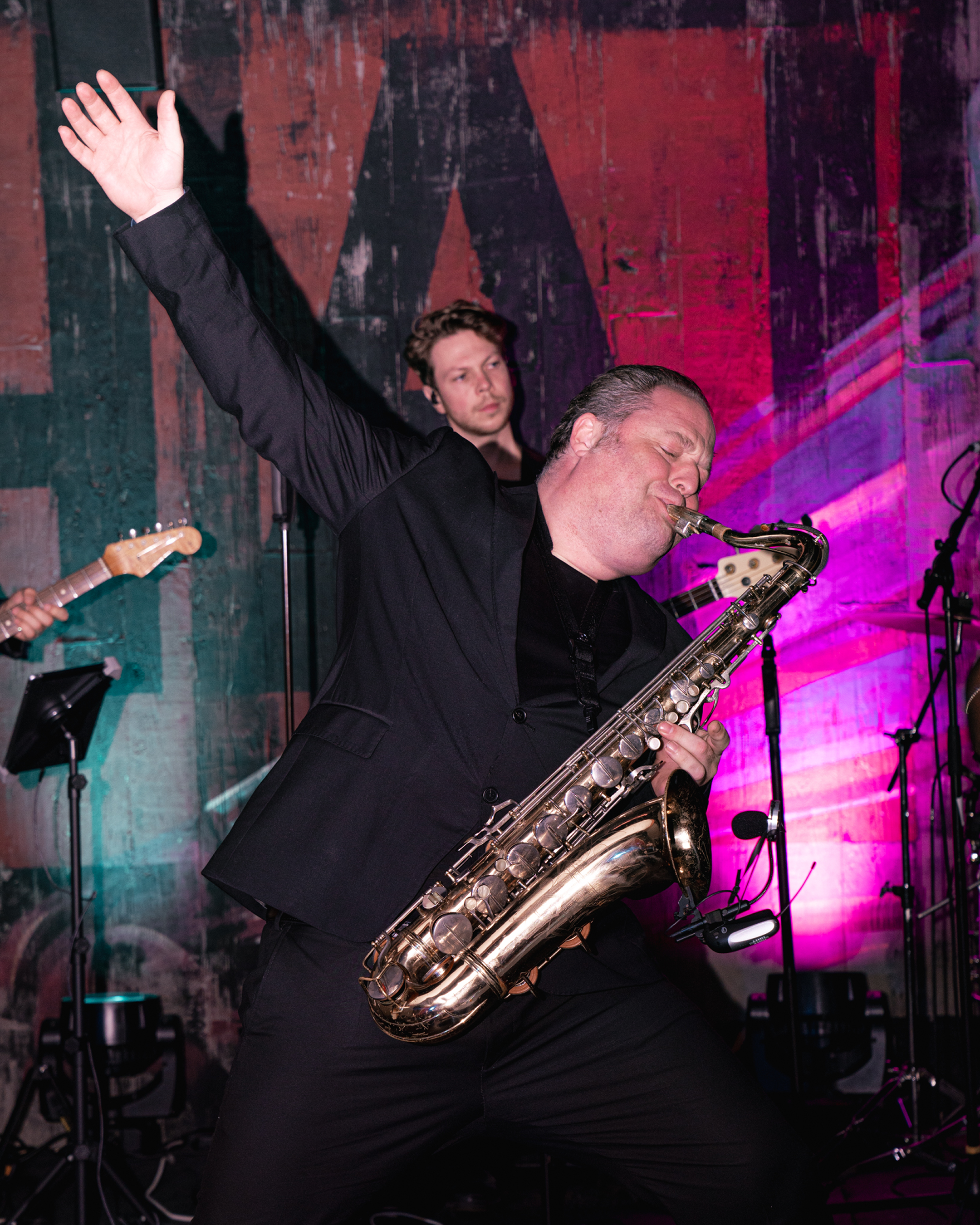 A man playing a saxophone passionately with his eyes closed and one arm raised, while another musician in the background watches, in a concert setting with colorful lighting.
