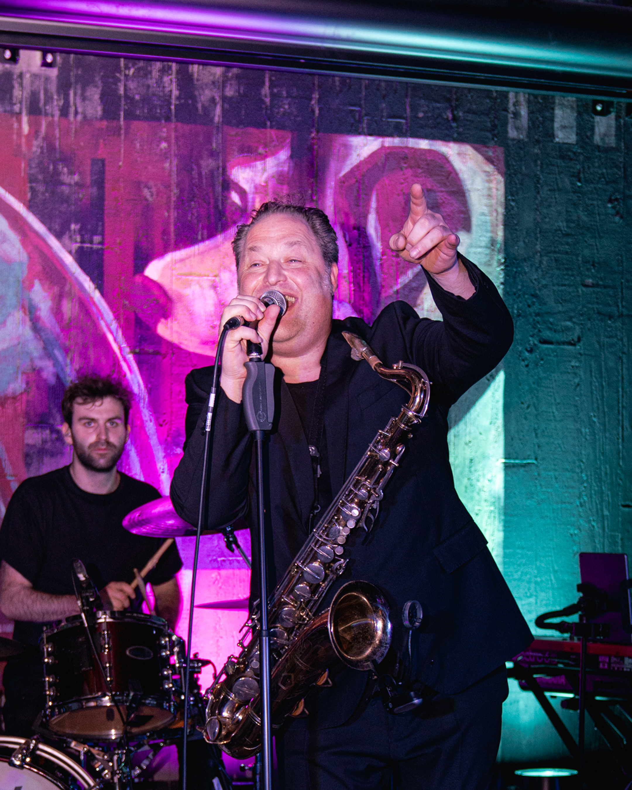 Man singing into a microphone on stage with a saxophone, with a drummer playing behind him, colorful stage lighting, and a textured brick wall in the background.