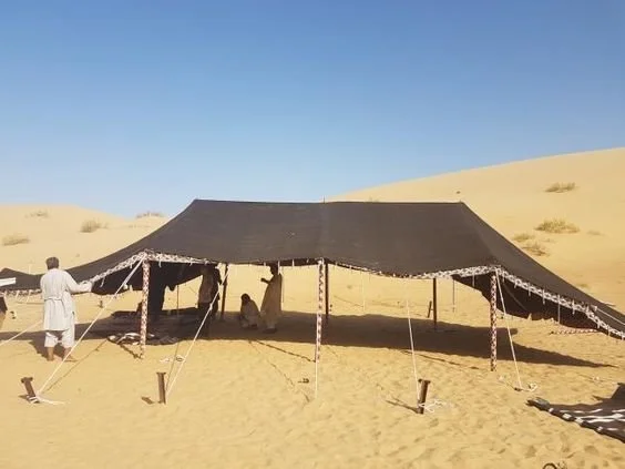 A large black canopy bedouin tent set up in a desert with sand dunes in the background, people underneath and around it.
