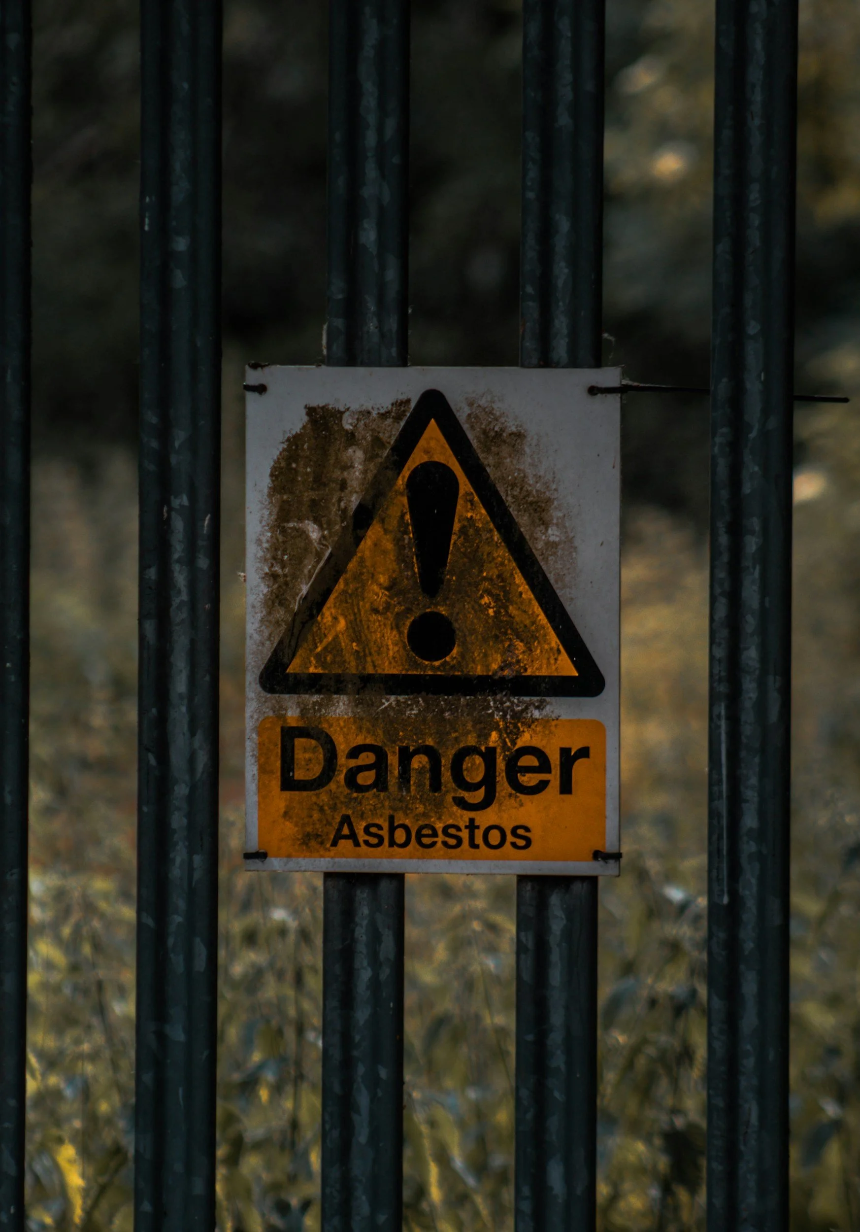 A metal fence with a yellow and black warning sign that says 'Danger Asbestos' in English and Spanish, indicating a hazardous area.
