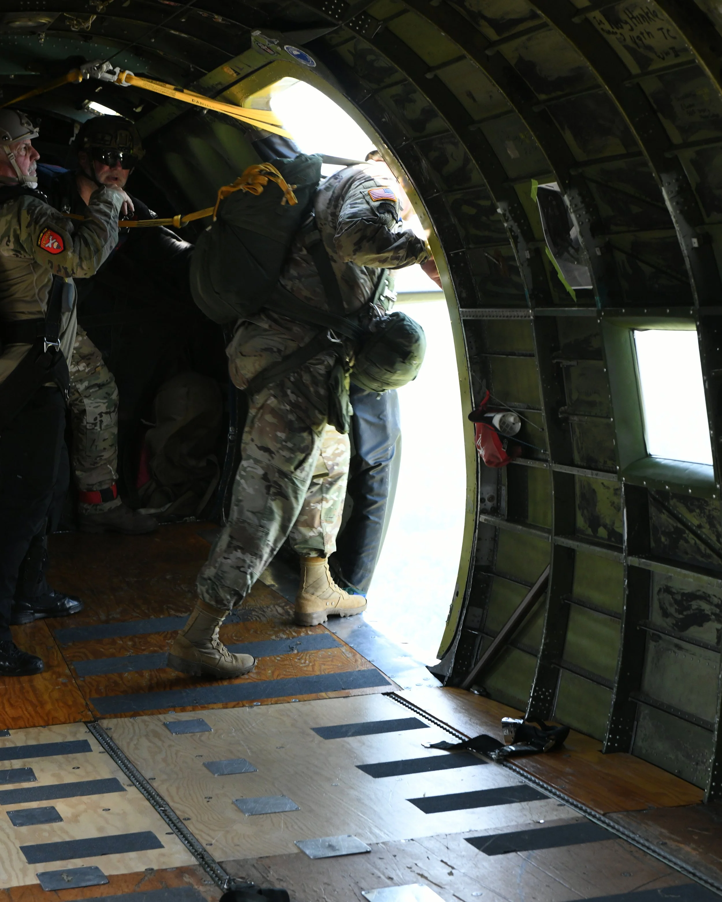 Military personnel preparing to exit a military aircraft through an open side door, wearing camouflage uniforms and tactical gear, with a bright sky outside.