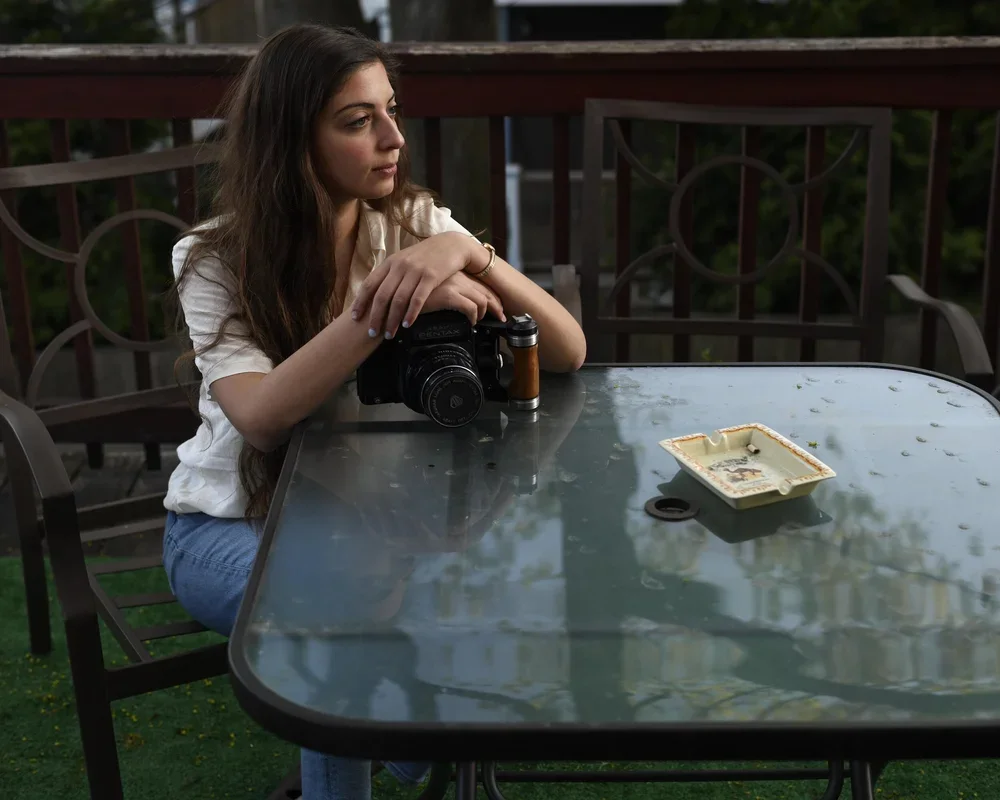 A woman with long brown hair sitting at an outdoor glass table, holding a camera with her arms resting on the table, looking contemplative. The table has an empty ashtray on it.