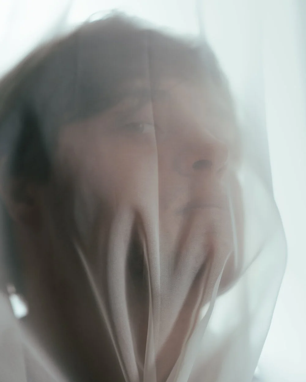 A person behind a sheer, semi-transparent curtain, with part of their face and hand visible through the fabric.