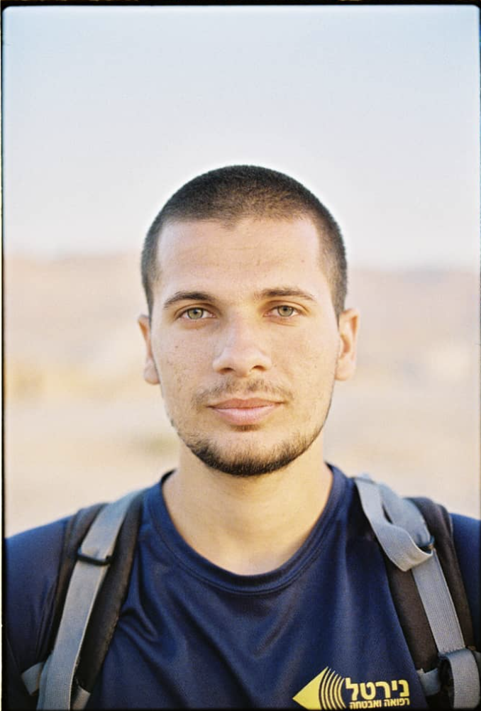 A young man with short brown hair, a light beard, and green eyes, wearing a navy blue T-shirt with a yellow logo, and carrying a gray backpack, standing outdoors in a desert landscape with a clear sky.