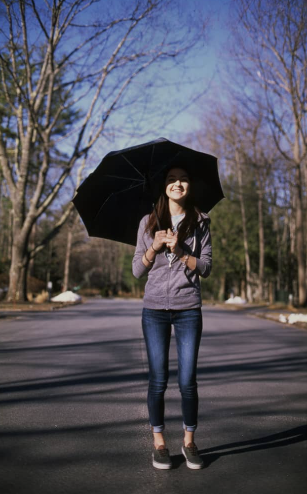 A young woman holding a black umbrella standing on an empty street in a park during winter, with leafless trees and a blue sky in the background, smiling.
