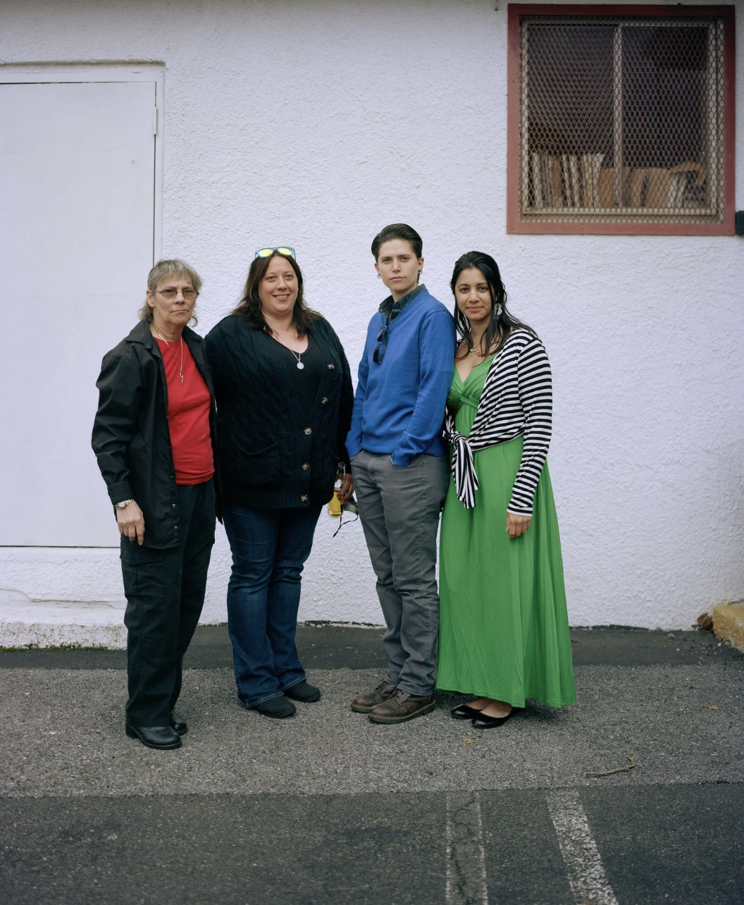 Four women standing side by side outdoors against a white textured wall, dressed in casual and semi-formal attire, with a window and a door behind them.