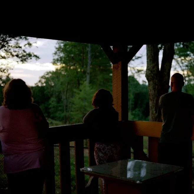 Three people standing on a porch, looking out at a wooded landscape during dusk.