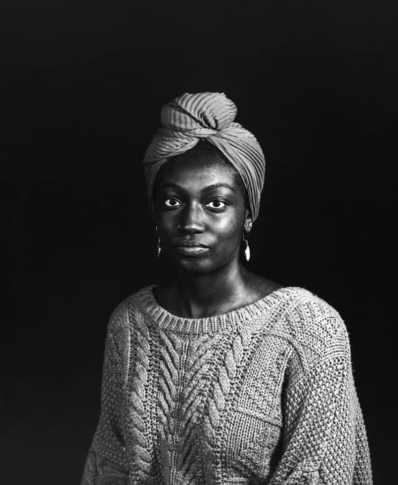 A black-and-white portrait of a woman with a headwrap, wearing a knitted sweater and earrings, against a dark background.
