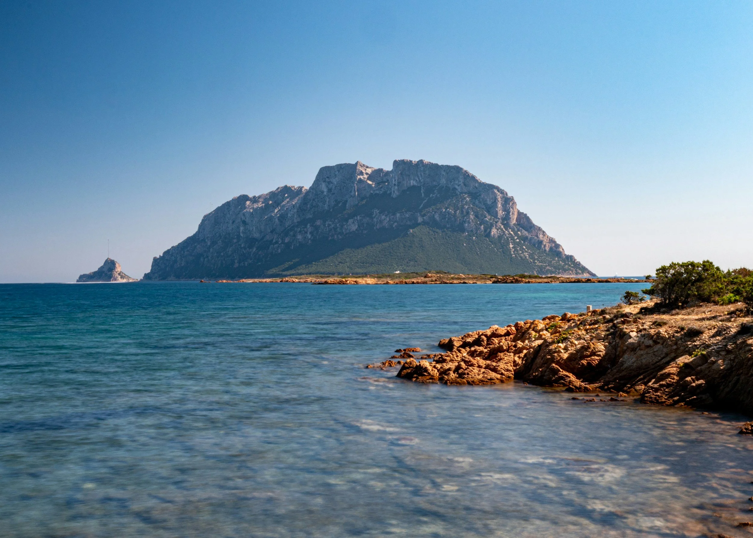 Coastal landscape with rocky shoreline and mountainous island in the distance.