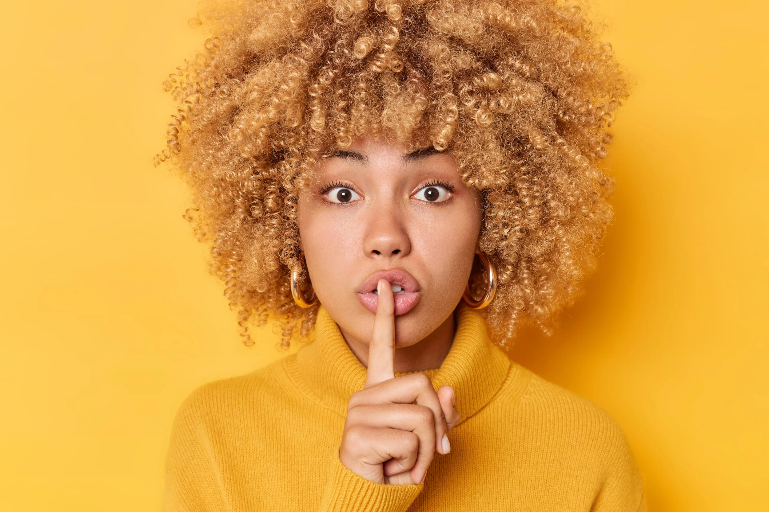 A curly haired black woman with her index finger pressed against her mouth with a shush gesture