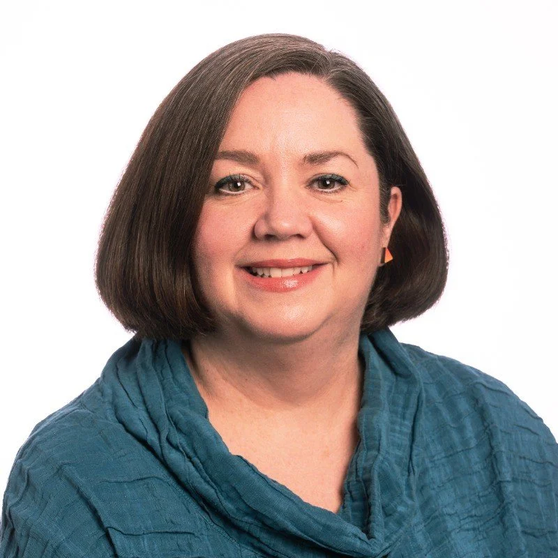 A middle-aged woman with shoulder-length brown hair wearing a blue top smiling against a plain white background.