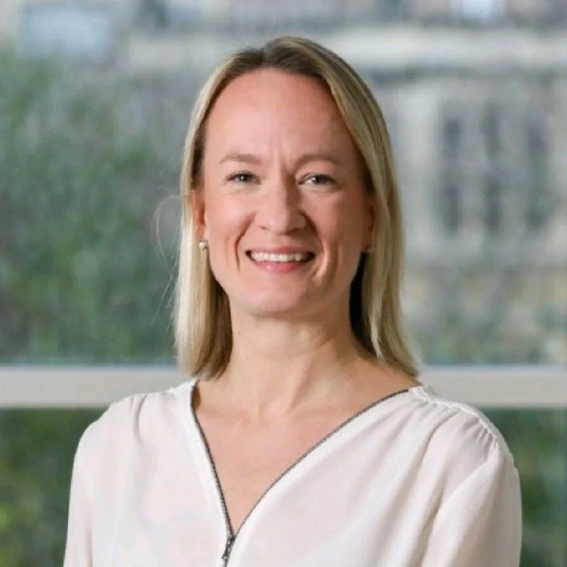 A woman with shoulder-length blonde hair smiling, wearing a white top, standing indoors with a blurred window and green trees in the background.