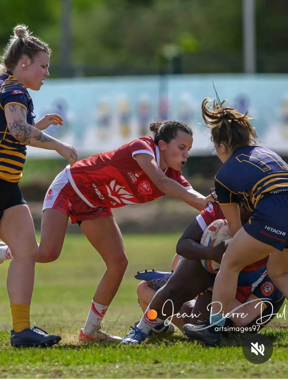 Jeunes femmes jouant au rugby, une en rouge essayant de prendre le ballon à une autre en bleu et jaune, lors d'un match en extérieur.