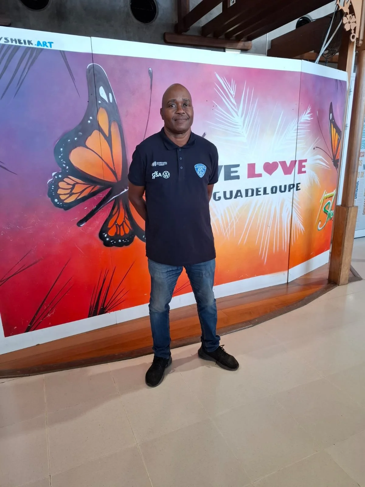 Un homme debout devant un panneau coloré avec une grande illustration de papillon et le texte "LOVE GUADELOUPE".