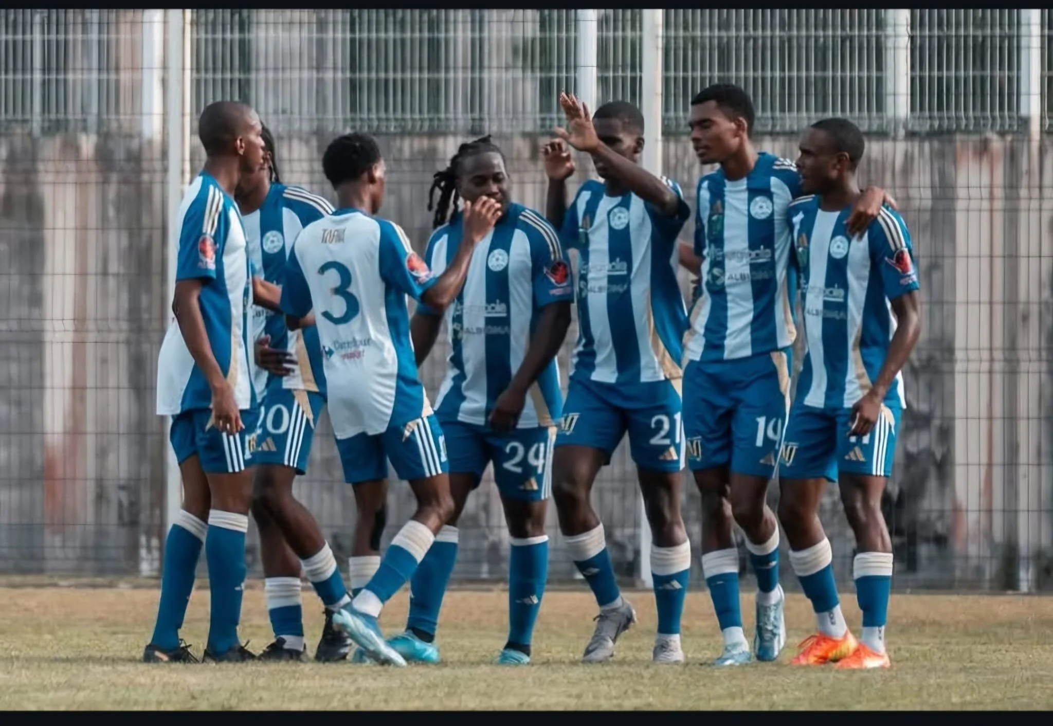 Groupe de joueurs de football en train de célébrer sur le terrain, portant des maillots et shorts bleus avec des rayures blanches, dans un stade extérieur.