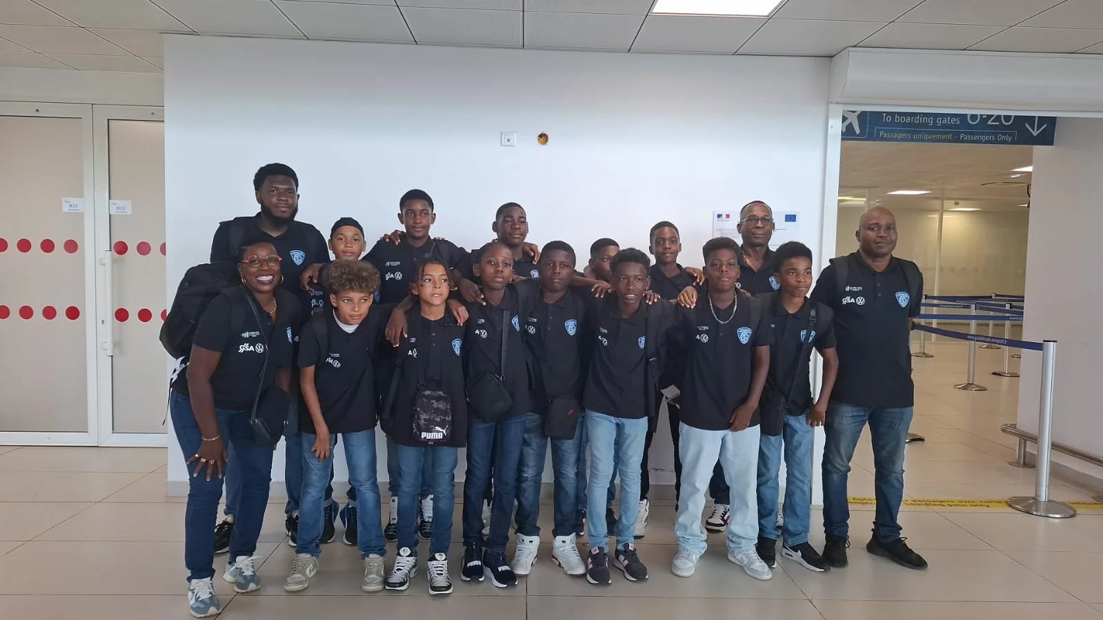 Groupe de jeunes et de deux adultes, tous portant des maillots noirs avec un logo, à l'aéroport près d'une porte d'embarquement.