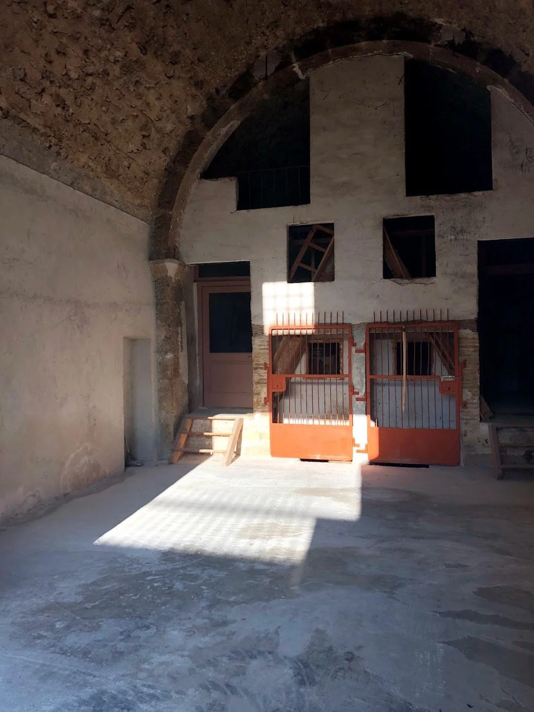 Indoor space resembling a historical or abandoned building with exposed brick ceiling, stone walls, and a concrete floor. There are small windows with wooden bars and a door with a small set of wooden stairs. Bright sunlight creates a shadow and illuminates part of the wall and floor.
