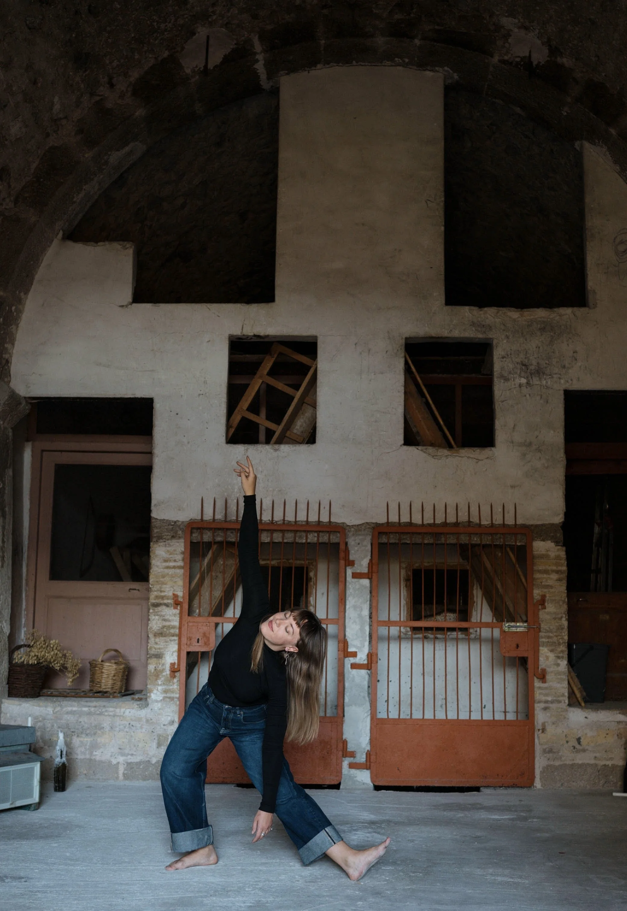 A woman with long brown hair, wearing a black long-sleeve top and wide-leg blue jeans, poses in front of an orange metal gate in a rustic interior with exposed brick and a painted white cross on the wall.