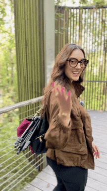A woman wearing glasses and a brown jacket, smiling on a balcony with a metal railing and green trees in the background.