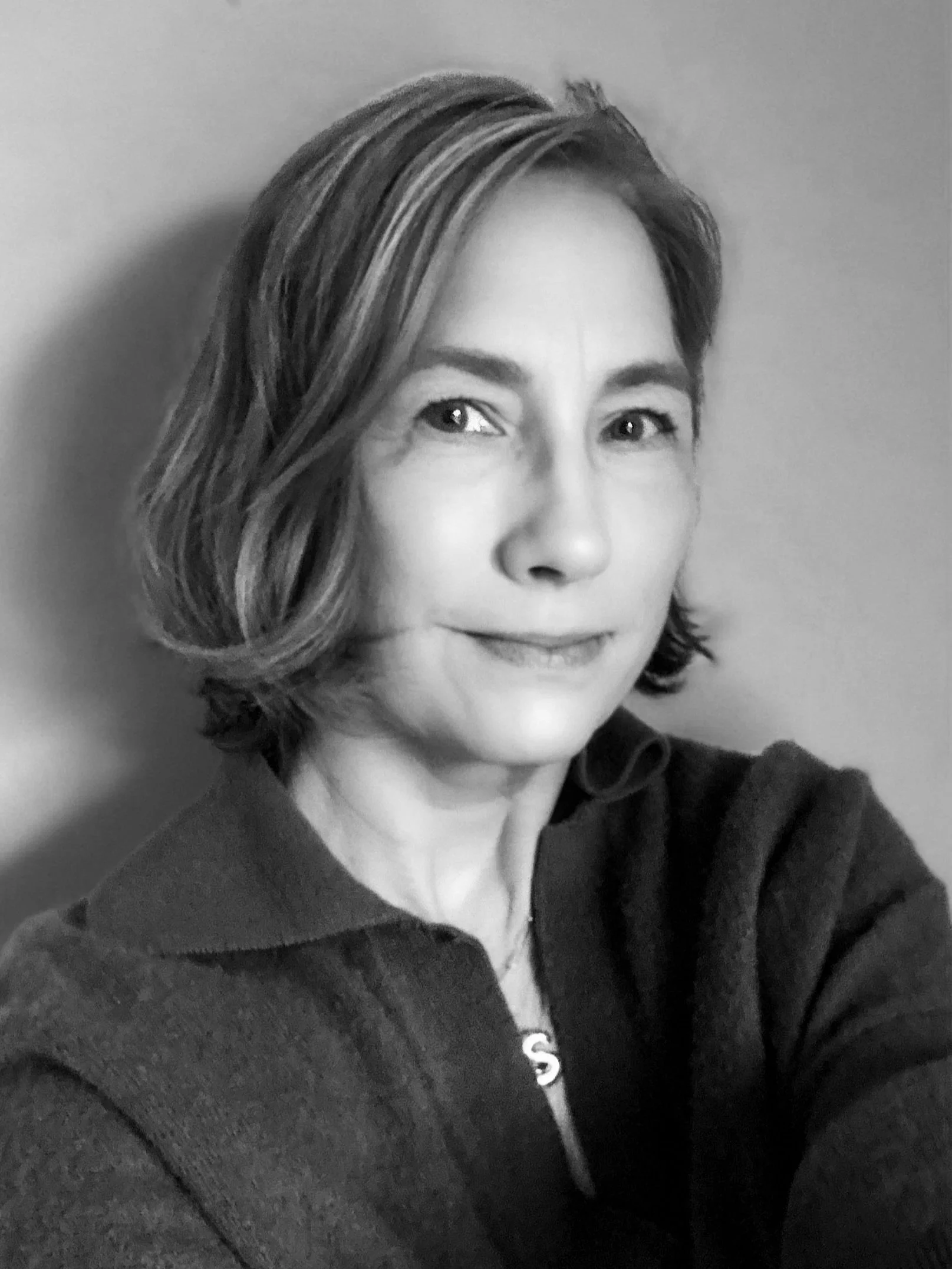 A woman with shoulder-length light brown hair and fair skin is wearing a maroon top and has a gentle smile, taking a selfie in front of a plain wall.