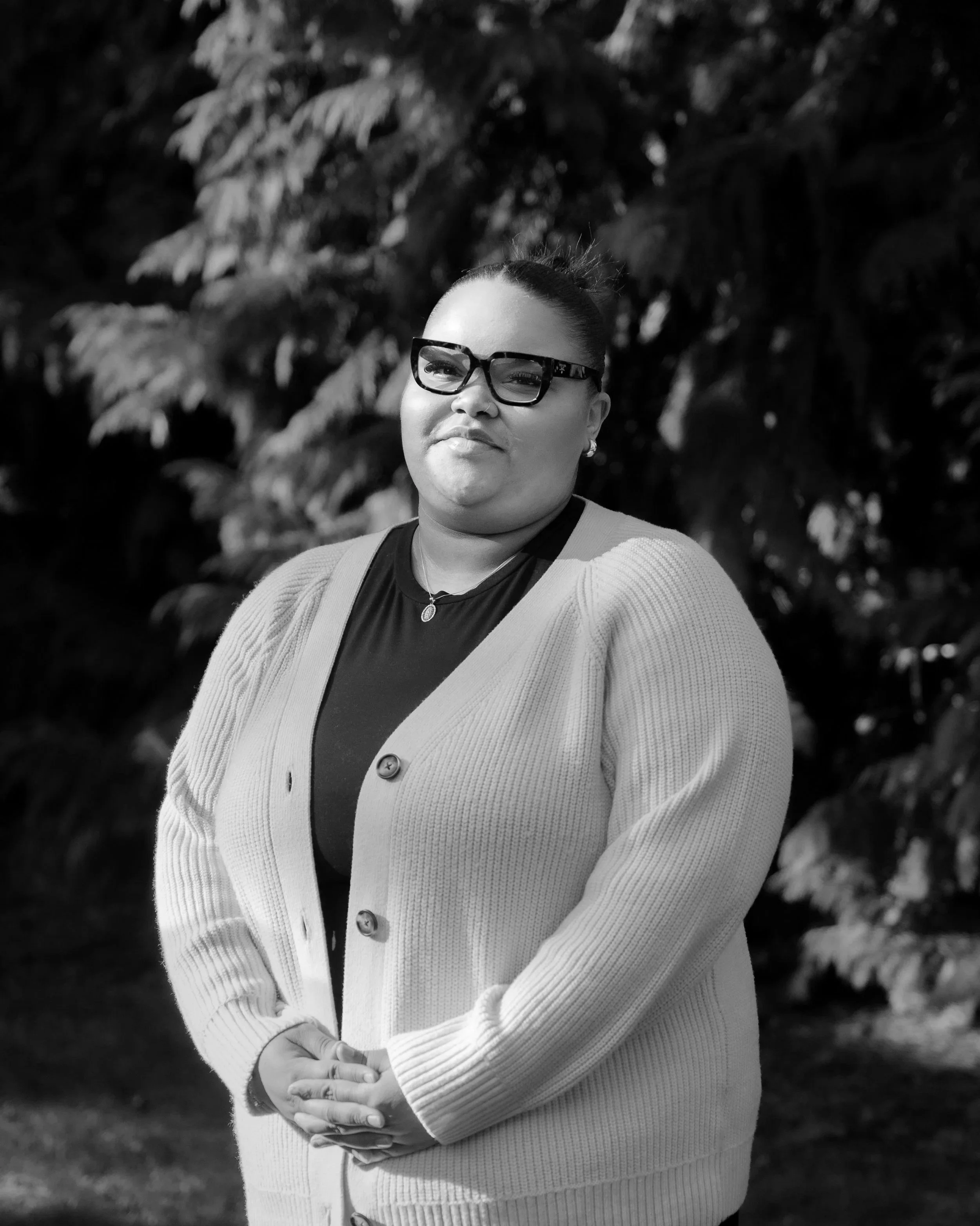 A woman standing outdoors with a background of trees, wearing glasses, a necklace, and a cardigan over a top, with her hands clasped in front of her.