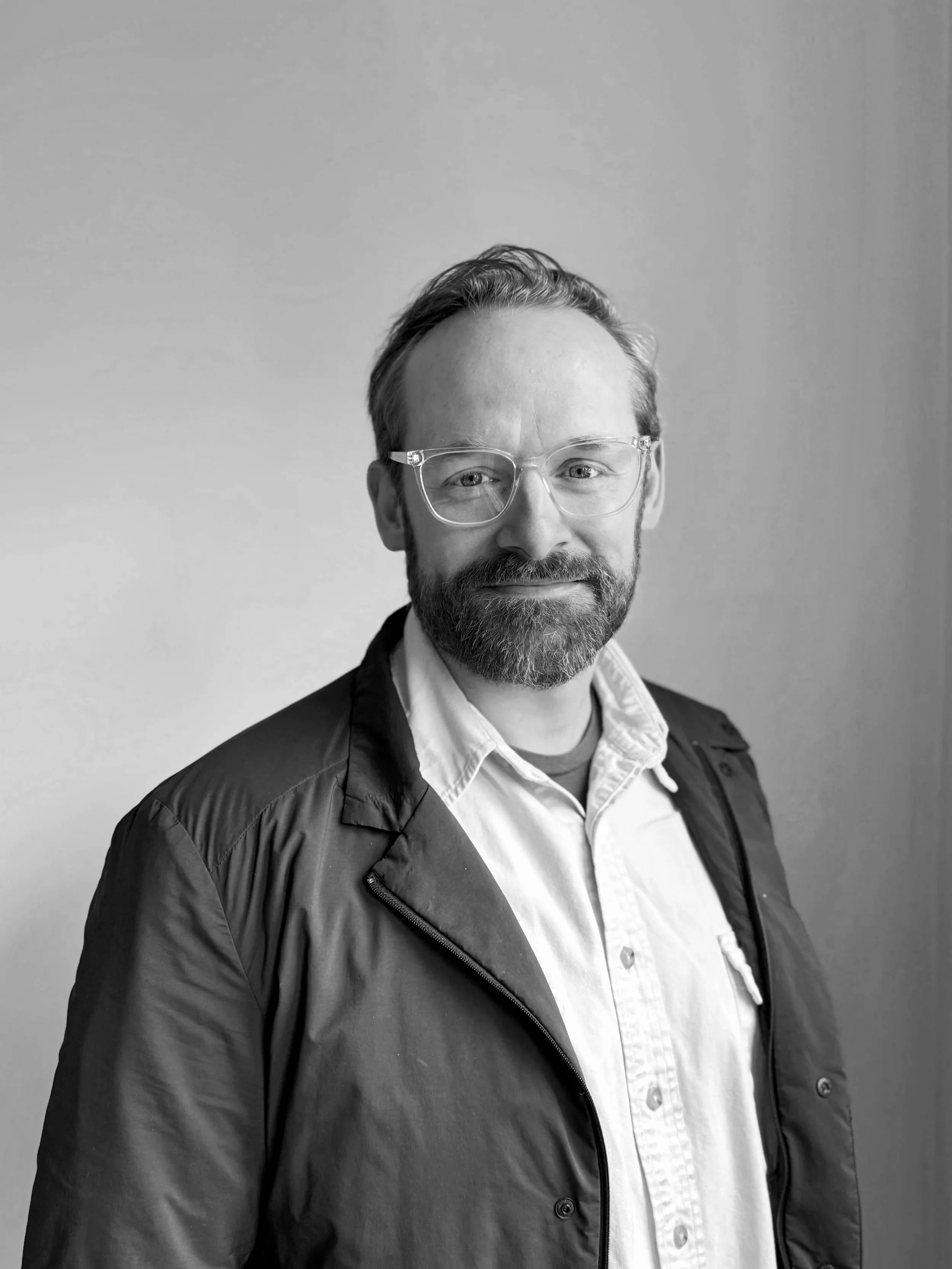 A man with red hair and beard wearing clear glasses, a white collared shirt, and a dark jacket standing against a plain light-colored wall.