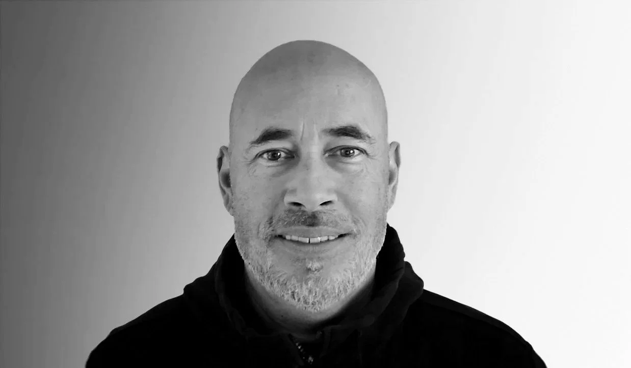 Portrait of a middle-aged man with a bald head, grey beard and mustache, wearing a black jacket, smiling, against a neutral background.