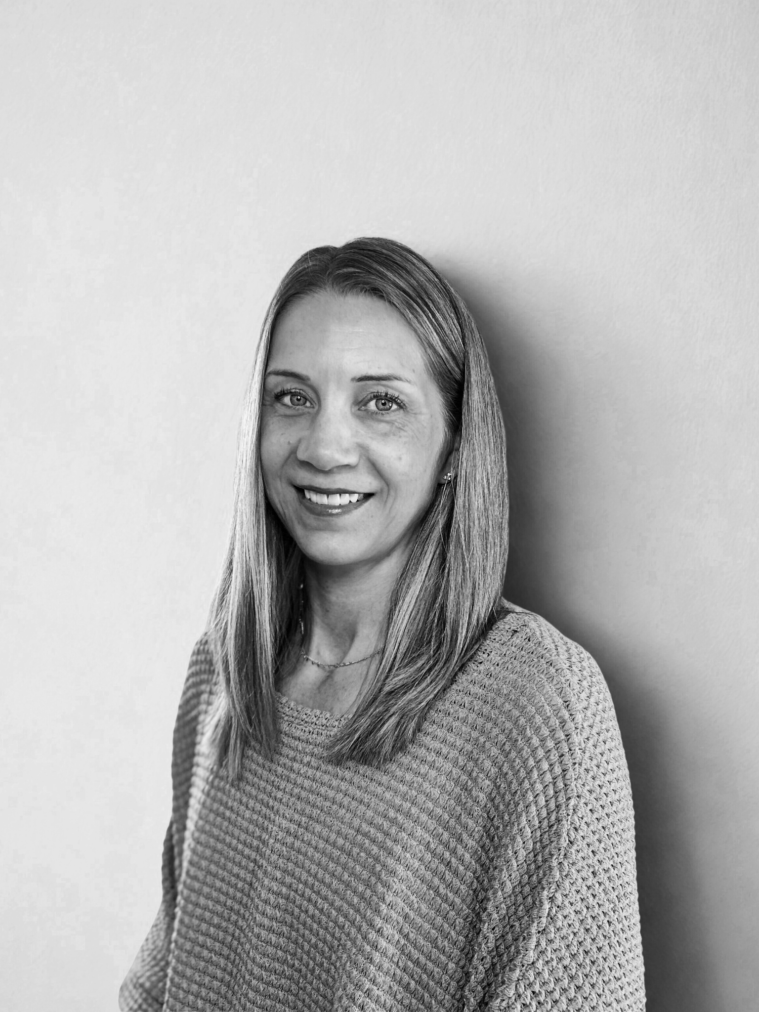 A woman with straight blonde hair, wearing a beige textured sweater, smiling and standing against a light blue wall.