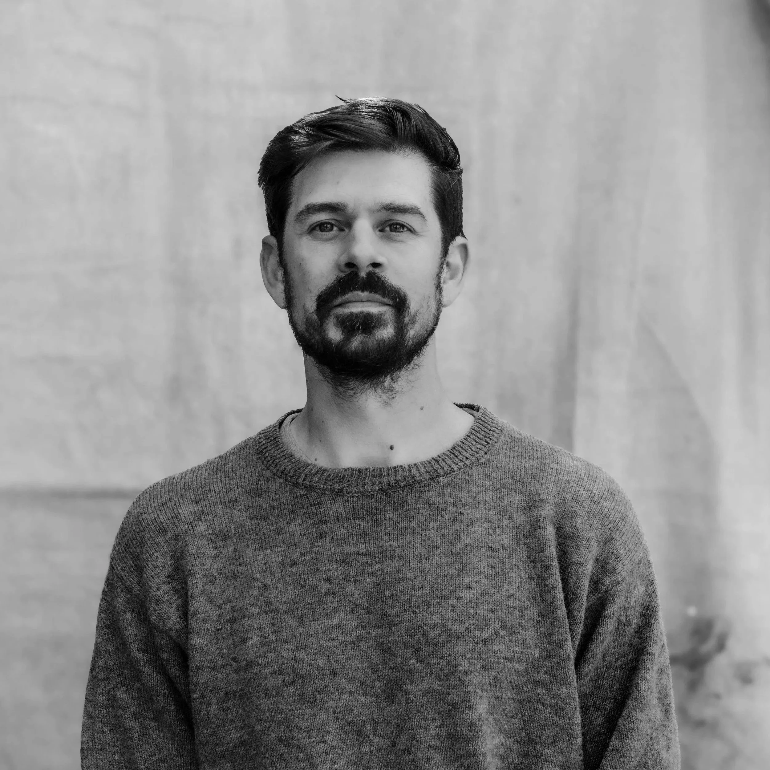A man with dark hair, a beard, and mustache wearing a blue sweater standing outdoors against a plain neutral background.