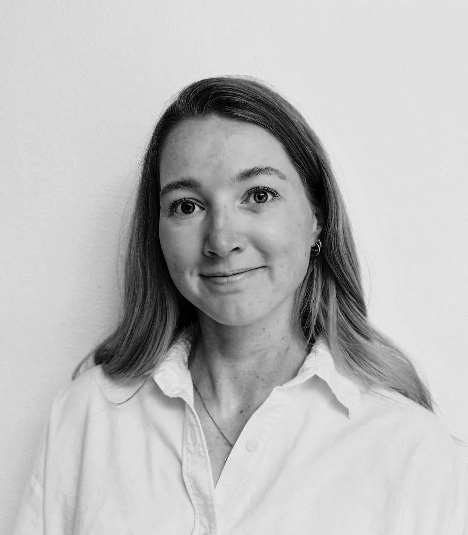 Portrait of a young woman with light skin, blue eyes, and light brown hair, wearing a white shirt, standing against a plain white wall.