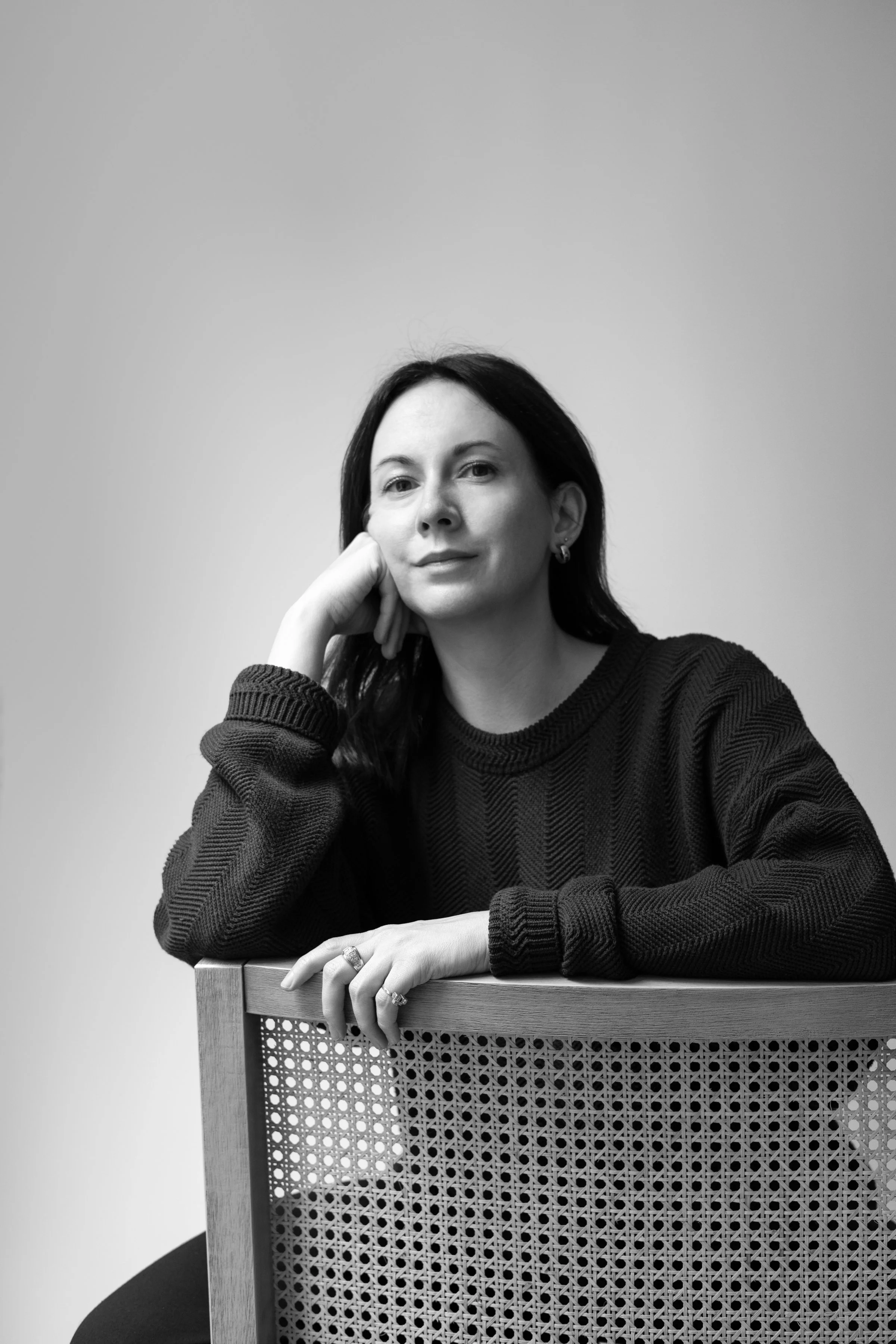 Black and white portrait of a woman with dark hair, wearing a sweater, resting her chin on her hand, sitting behind a wooden chair with a woven backrest, against a plain background.