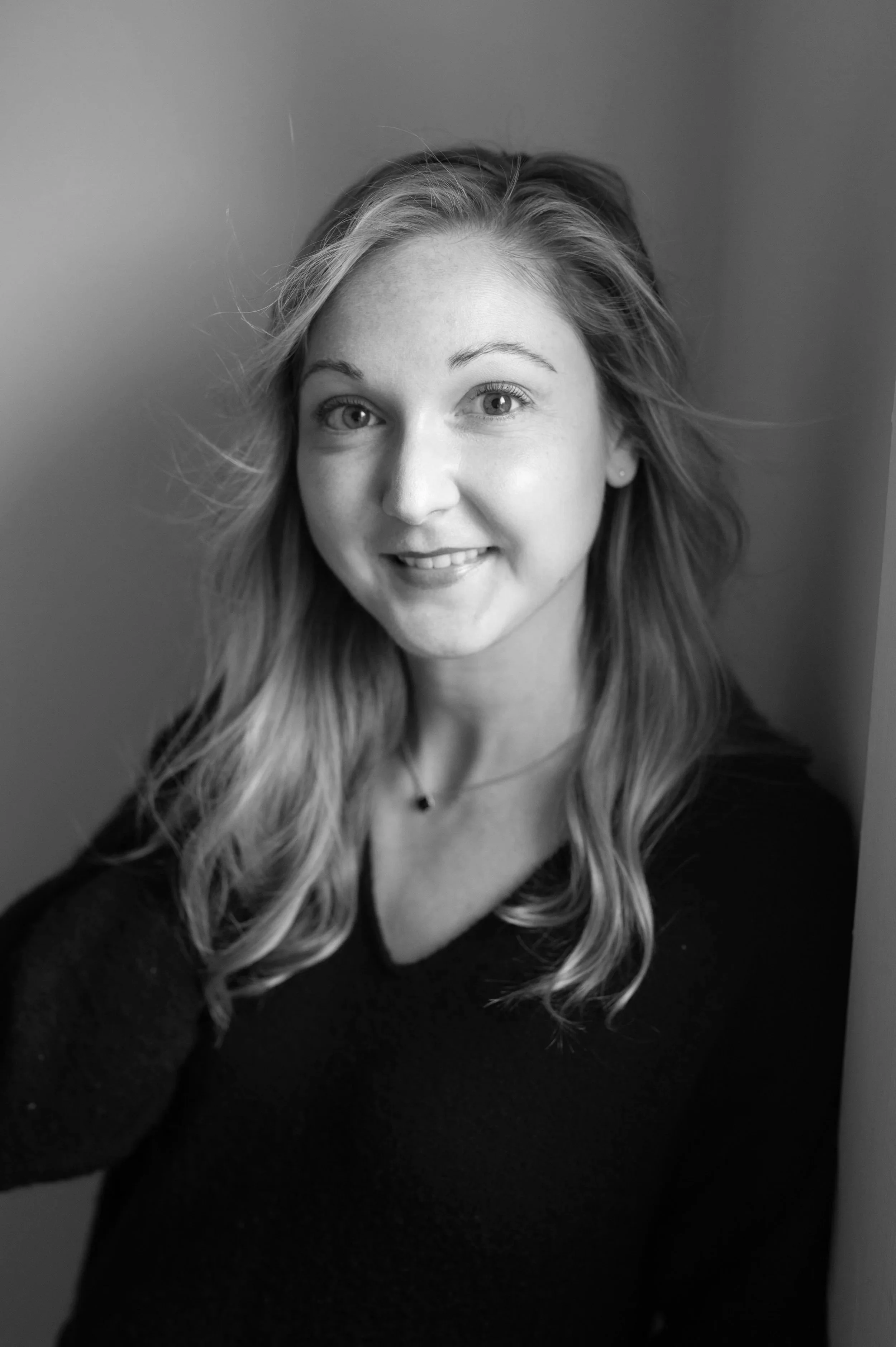 Portrait of a young woman with long blonde hair, wearing a black top, smiling, and standing against a neutral background.