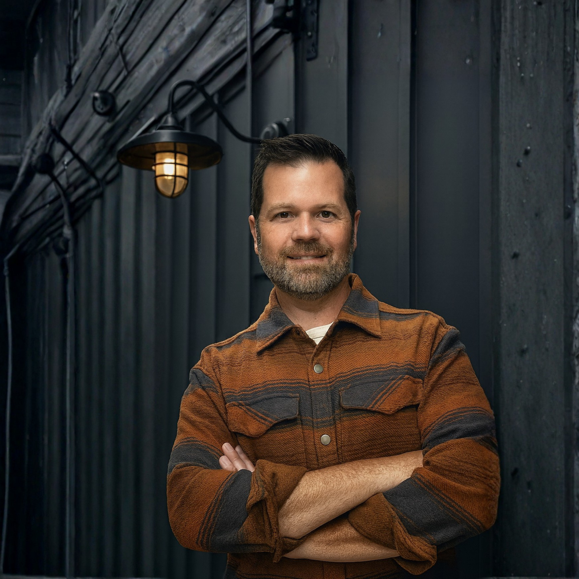 man standing with arms crossed in striped orange/grey shirt jacket against a grey wall and a barn light casting a soft glow