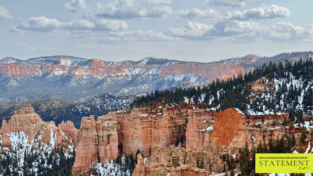 View of Bryce Canyon National Park with snow-capped rugged red rock formations, forested hills, and a cloudy sky.