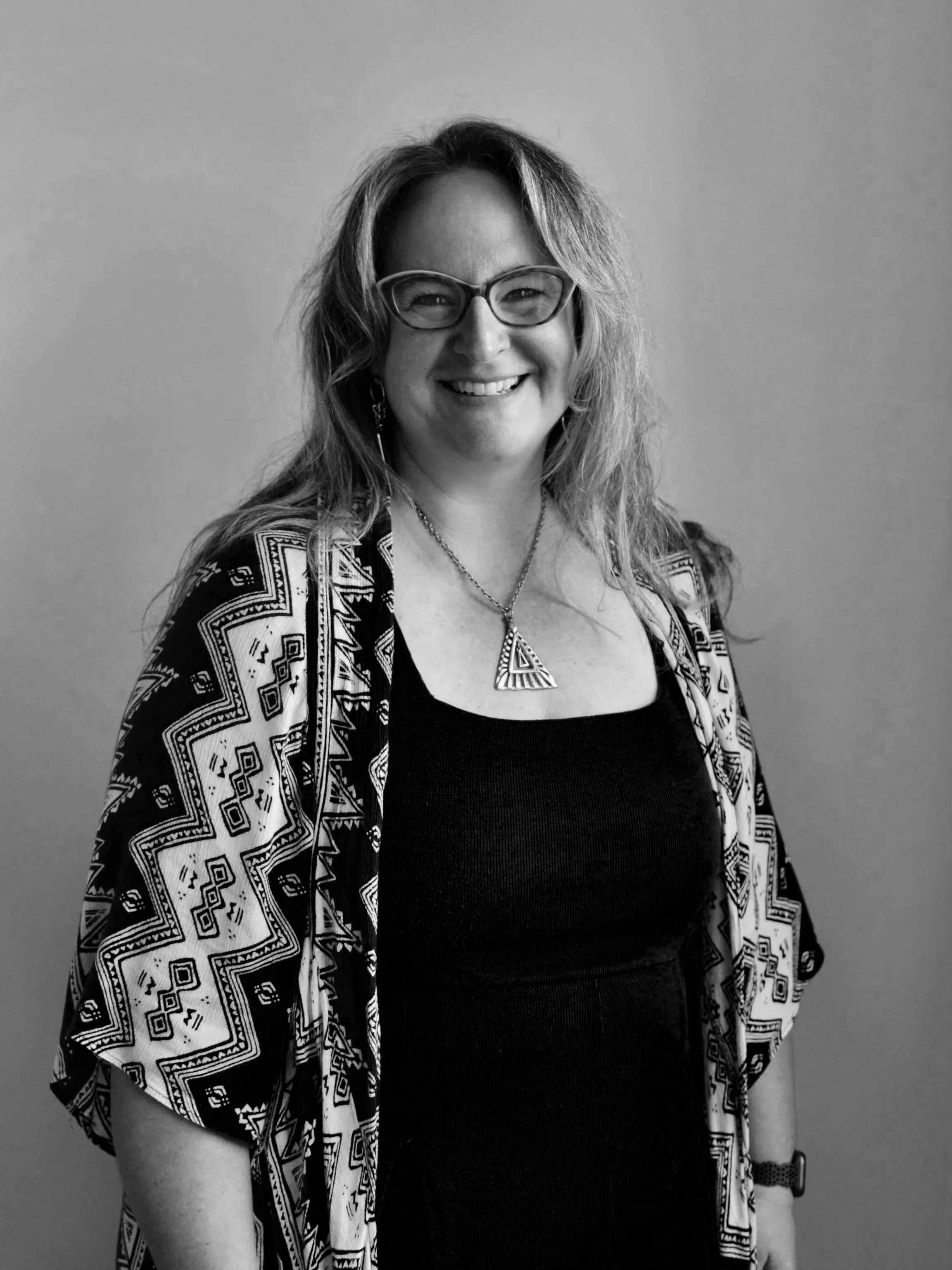 A woman with blonde hair, glasses, and earrings, wearing a blue dress and a patterned black and white shawl, smiling and standing against a plain wall.