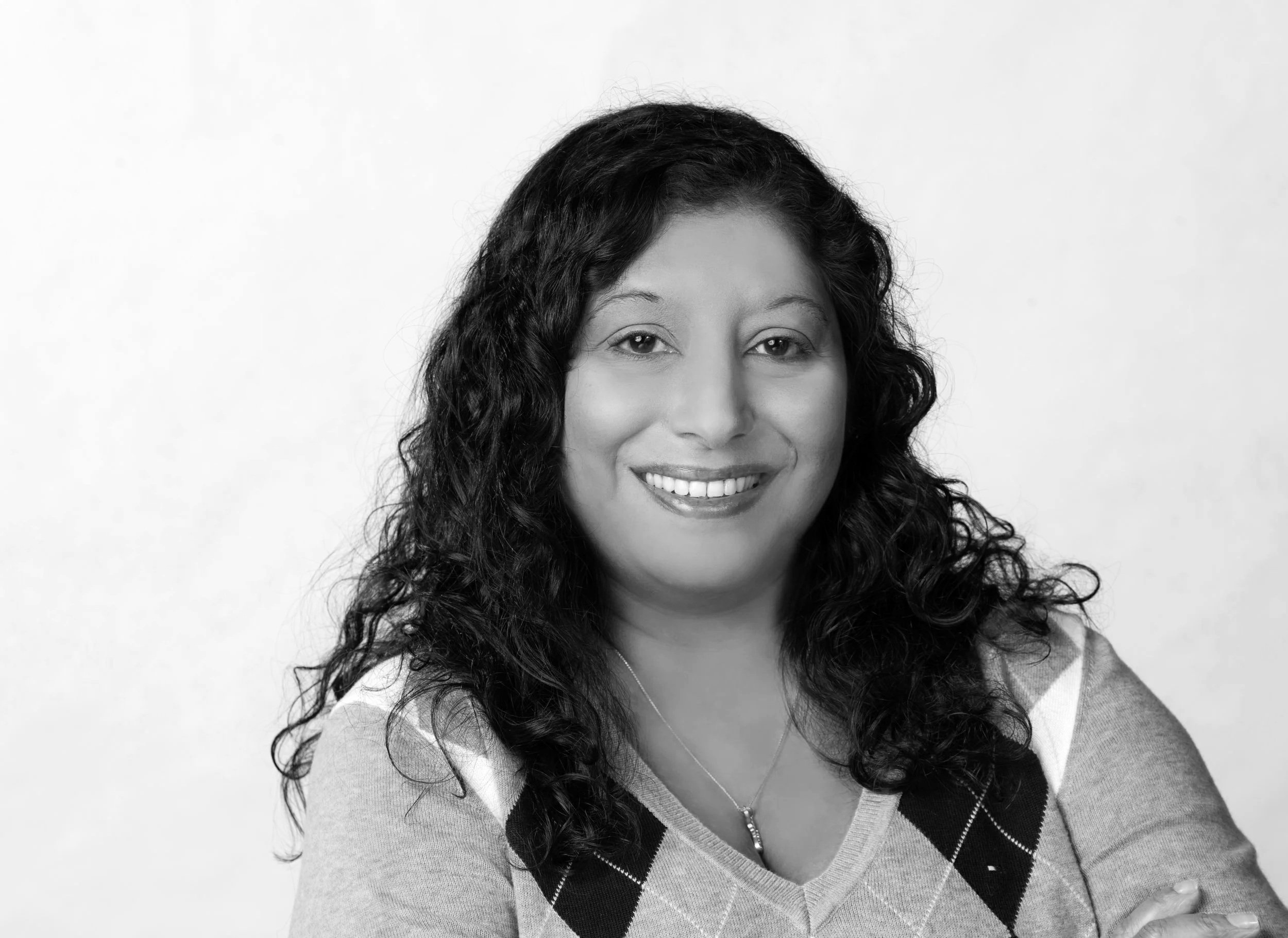 Portrait of a woman with long, curly dark hair smiling, wearing a blue argyle sweater and silver necklace, crossed arms, against a light gray background.