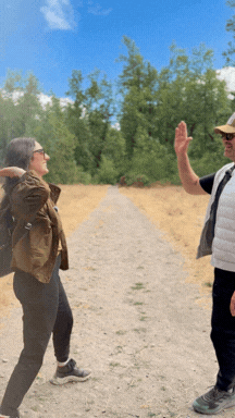 Two people standing on a dirt trail outdoors, greeting each other with a high-five, surrounded by trees and a blue sky.
