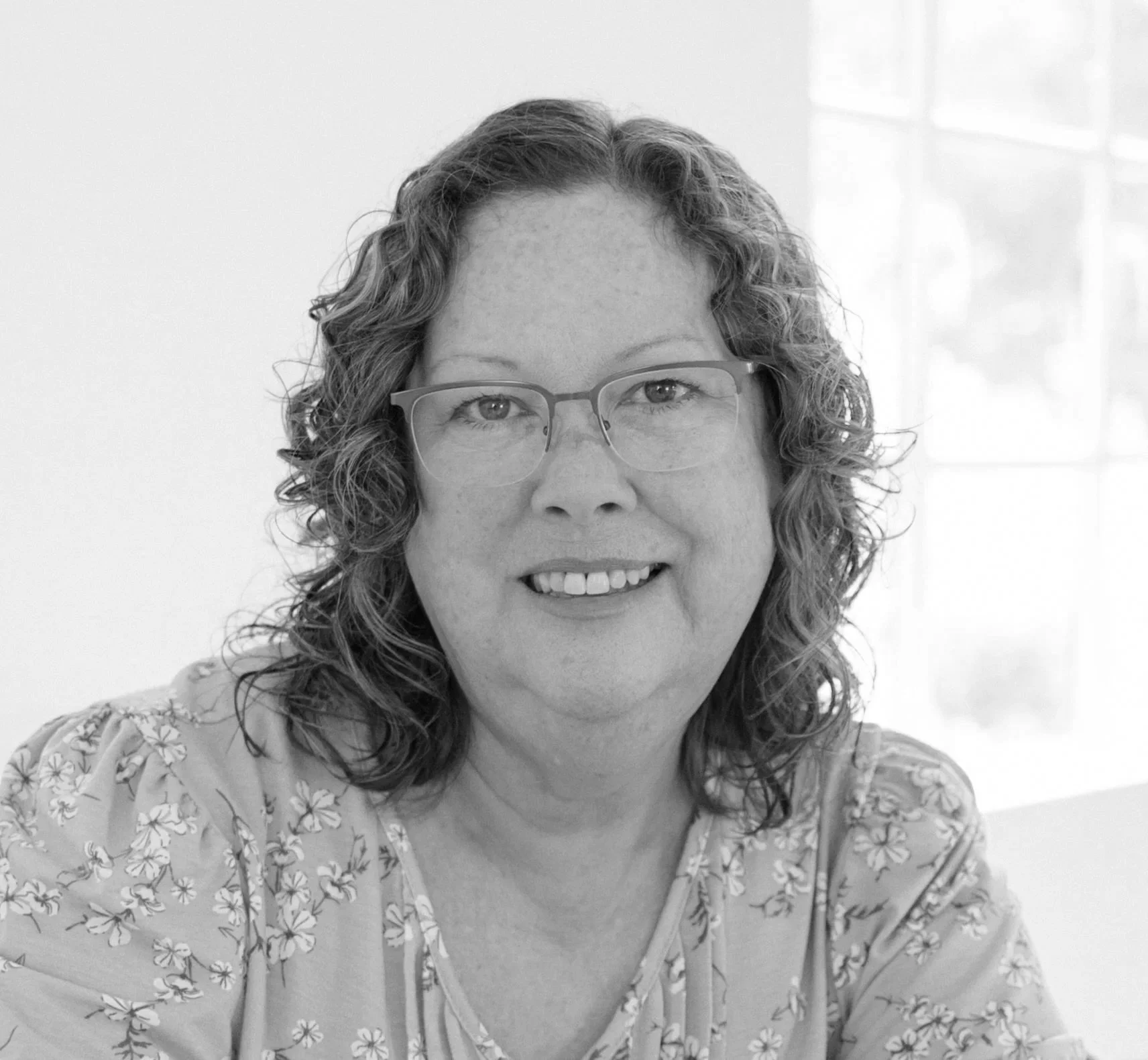 A woman with curly hair and glasses smiling, wearing a floral-patterned top, sitting in front of a window.