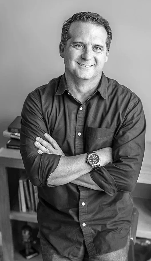 A smiling man with short brown hair, wearing a dark blue button-up shirt with sleeves rolled up, standing with arms crossed in an office space.