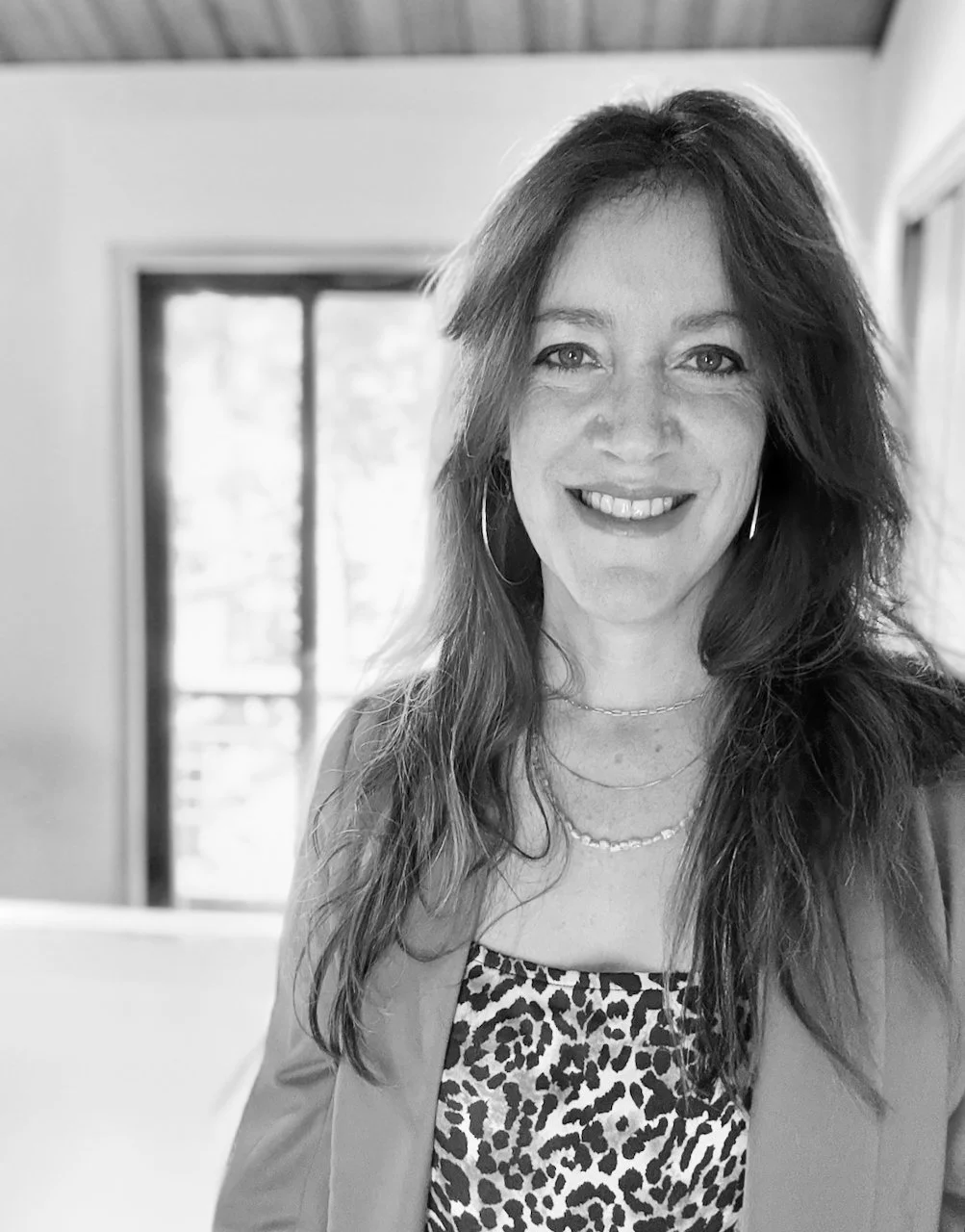 Black-and-white photo of a woman smiling, with wavy hair, wearing hoop earrings, layered necklaces, patterned top, and a blazer, standing near a window.