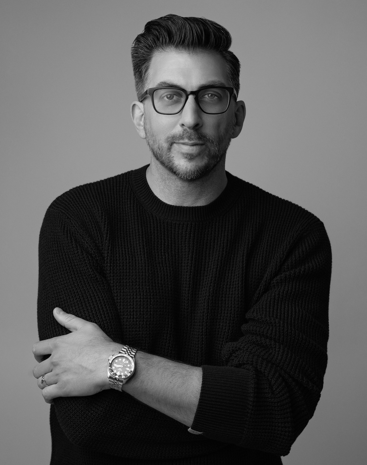 A man with dark hair, beard, and glasses wearing a black knit sweater, standing against a plain gray background, with arms crossed and a silver watch on his left wrist.