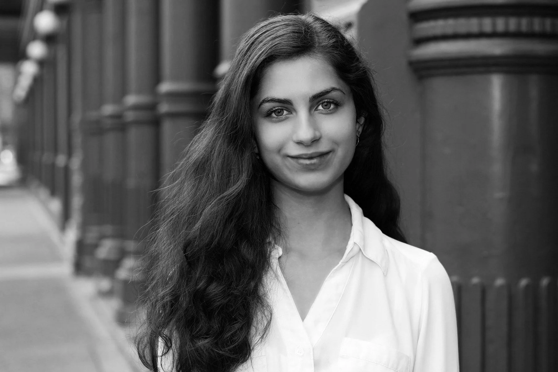 Black and white portrait of a young woman with long wavy hair, wearing a white shirt, standing outdoors near a brick building with large columns.