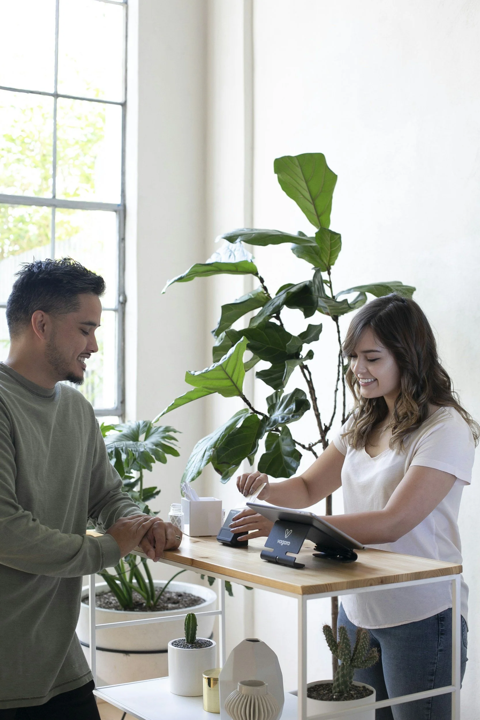 A man and a woman are standing at a wooden counter in a store with houseplants in the background. The woman is smiling and showing a product to the man, who is smiling and leaning slightly forward.