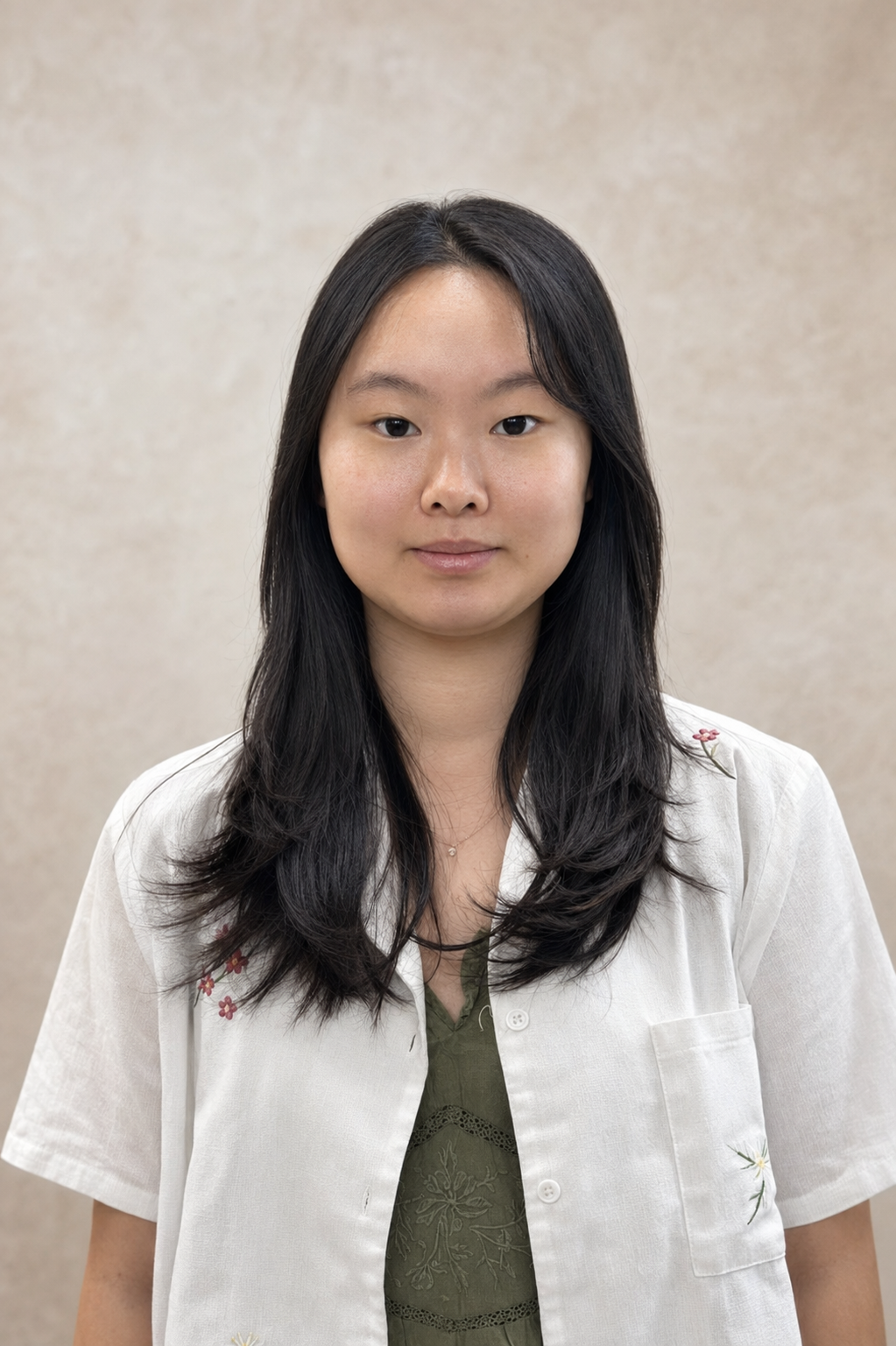 A young woman with long black hair and a calm expression, wearing a white short-sleeved shirt over a green top, standing against a plain beige background.
