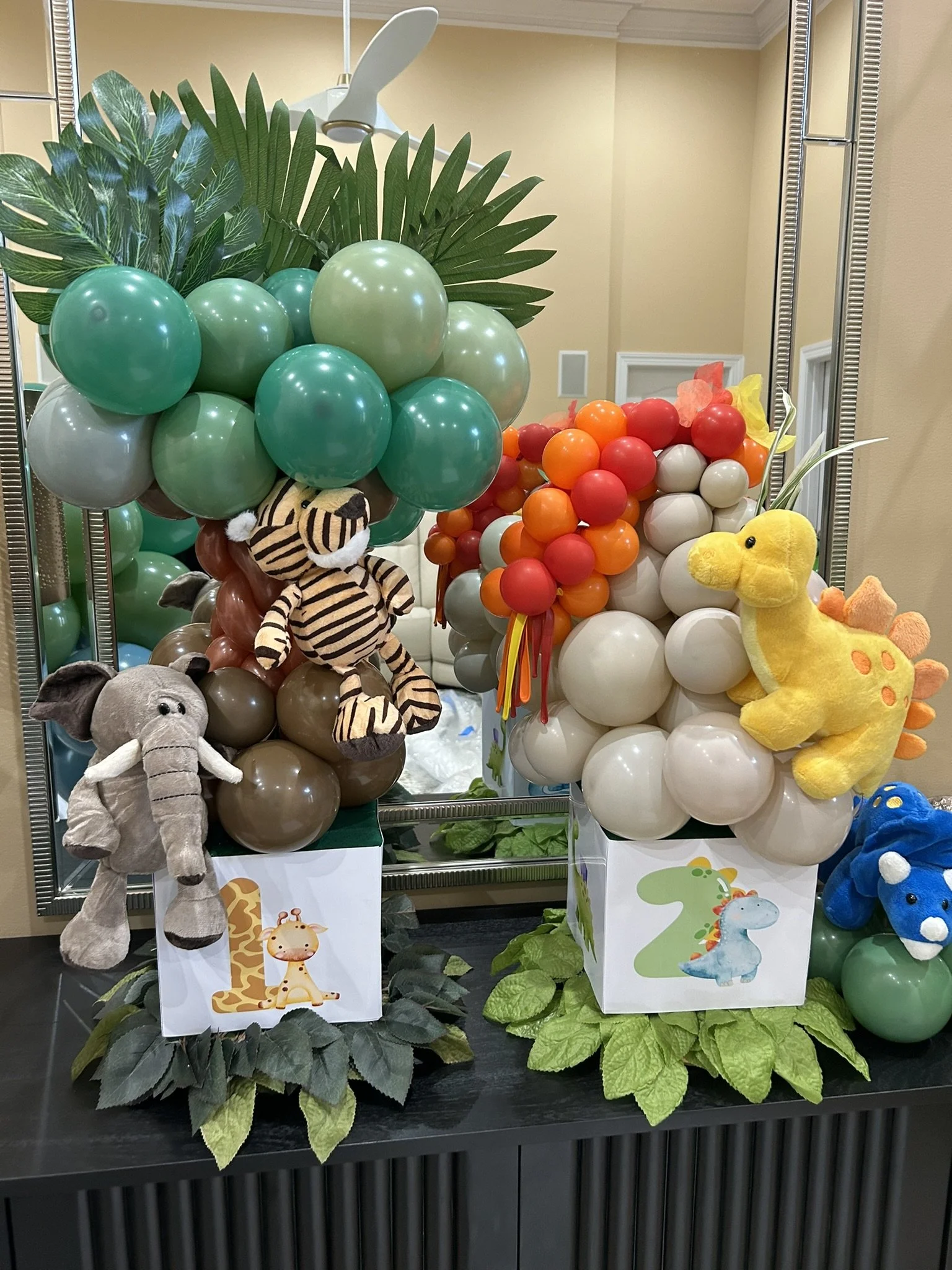Colorful balloon decorations featuring animal plush toys including a zebra, giraffe, elephant, dinosaur, and a yellow duck, with green artificial plants and a mirror in the background.