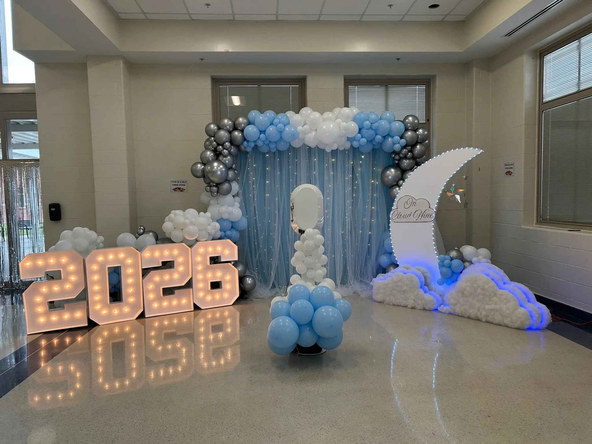 Decorative setup with white, light blue, and silver balloons arranged in an arch and clouds, with a large illuminated 